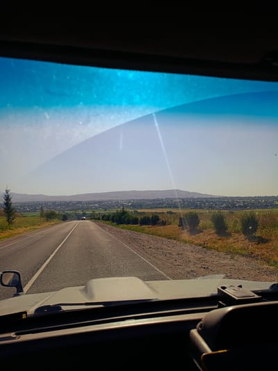 The image is a point-of-view shot from inside a vehicle looking out through the windshield at a road leading towards a distant town. The road is paved with a white line dividing the lanes and is bordered by dry, grassy terrain. In the distance, a town is visible nestled among rolling hills under a clear, hazy sky. The foreground shows the dashboard and a side mirror of the vehicle. The time of day appears to be daytime, with the sun casting shadows and creating lens flare on the windshield. The environment is rural and appears to be arid or semi-arid.