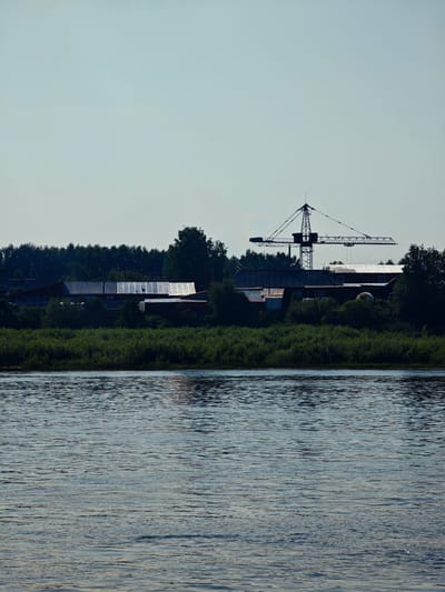 This is a wide shot of a body of water with a village and a construction crane on the opposite bank, located in the village of Verkhne-Pashino, Russia. The water is calm with gentle ripples, reflecting the sky and the bank. The bank is lined with dense green foliage and trees. Behind the foliage, several buildings are visible, including what appears to be a long, corrugated metal-roofed structure and other houses. A large construction crane stands prominently behind the buildings, its silhouette stark against the clear, pale blue sky. The overall scene suggests a rural or semi-industrial area situated near a significant body of water. No people are visible in the image.