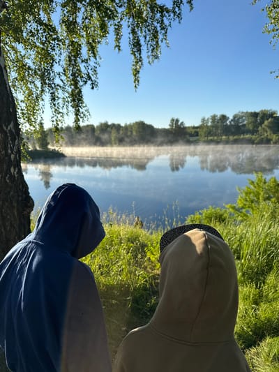 Two individuals, seen from behind, are observing a tranquil river scene. They are positioned near the bank, with tall grass and a tree to their left. The river is calm, reflecting the clear blue sky and the lush green trees on the opposite bank. A gentle mist hovers over the water's surface, illuminated by the morning sun. The overall atmosphere is serene and peaceful. It is not possible to determine the specific location within Beryozovka, Russia from the image.