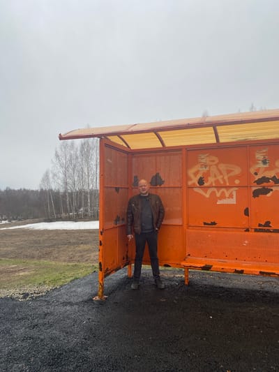 A man stands within an orange bus shelter in what appears to be Буль-Кайпаново, Russia. The shelter features graffiti and some rust. The man wears a brown jacket and dark trousers. Outside the shelter, the ground is paved on one side and transitioning to a field with snow and bare trees on the other. The sky is overcast.