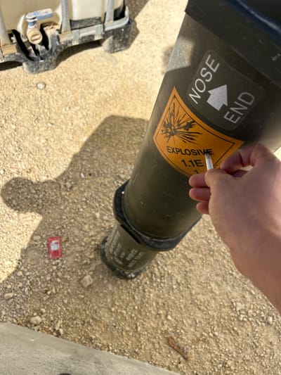 A person holds a cigarette to an explosive ordnance. The device is olive green with a yellow label marked "EXPLOSIVE 1.1E," and an arrow indicating the "NOSE END." The device sits on dirt with small rocks. A red object is near the base of the device, with a section of a white container and its metal frame.