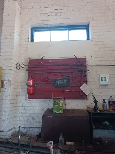 The image depicts an interior scene, likely a workshop or storage area, located in Vuhlehirsk, Ukraine. The walls are constructed of white-painted brick. A window with dark framing is visible in the upper portion of the image. Dominating the center of the frame is a large, red wooden panel mounted on the wall, which serves as a rack for various firefighting tools. These tools include axes, a fire hook with a coiled handle, and other implements. A red fire extinguisher is mounted to the left of the panel. Below the panel, a dark sign reads "ПОСЫПОЧНЫЙ МАТЕРИАЛ," which translates to "Sprinkling Material." To the right of the red panel, a small white first-aid kit with a green cross is mounted on the wall.
In the foreground, a large, dark brown metal cabinet or chest is visible. A box for a "PRO CRAFT Brush Cutter T5600" is placed on top of this cabinet. Various tools and items are scattered on a workbench in front of the cabinet, including a blue plastic bottle of liquid, a power tool with a saw attachment, and other miscellaneous equipment. A portion of a grey metal shelving unit is visible to the right, containing containers and other items. The floor appears to be concrete or a similar industrial material, with debris and coiled wires present. No individuals are clearly visible in the scene. The overall impression is one of a functional, somewhat cluttered, utilitarian space.