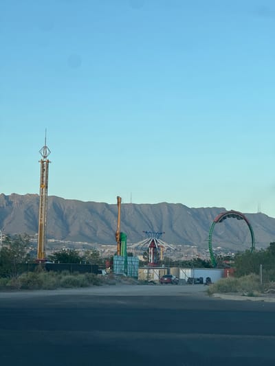The image depicts an outdoor scene with an amusement park in the foreground and a mountain range in the background. Several amusement rides are visible, including a tall drop tower on the left, a large circular ride with carriages in the middle, and a green roller coaster loop on the right. The area around the rides appears to be under construction or in a state of disrepair, with fencing, trailers, and debris present. The sky is clear and blue, suggesting a daytime setting. The mountain range in the background is rocky and arid. The foreground shows a paved surface, likely a road or parking lot, with some dry brush and vegetation. This scene is consistent with a location in or around Sunland Park, New Mexico, known for its desert landscape and proximity to mountains.
