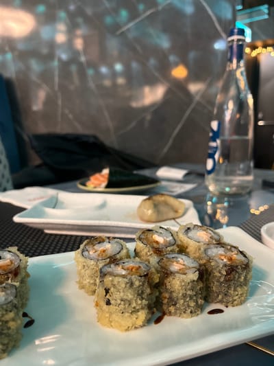 The image, rotated 90 degrees clockwise, presents an indoor dining scene, likely in a restaurant or bar in Basiglio, Italy. The primary focus is a long white rectangular plate filled with several pieces of deep-fried sushi rolls, also known as tempura rolls. These rolls are coated in a golden-brown, crispy batter, with visible fillings of rice, seaweed, and an orange-hued ingredient, possibly salmon, along with a creamy white substance, potentially cream cheese. Drops of a dark sauce, resembling soy or eel sauce, are present on the plate and rolls.

To the left of the main sushi plate, there is a smaller white dish holding what appears to be a single oyster or mussel. Further to the left, a clear glass bottle with a blue and white label stands upright on a dark, polished table with light, marble-like veining. Reflections of blurred, bright lights from the surroundings are visible on the bottle and the table surface, suggesting an evening or nighttime setting. Some indistinct objects and reflections are visible in the background, out of focus. The text on the bottle's label is partially obscured and too blurry to be clearly legible. No people are visible in the frame. The scene captures a moment during a meal, emphasizing the prepared food items.