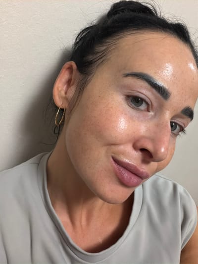 A close-up, head-and-shoulders shot shows a woman with dark hair pulled back, wearing a light gray top. She is leaning her head to her left, against a plain, textured off-white wall. The lighting highlights her dewy skin and prominent freckles. She has a gold hoop earring in her left ear and her eyebrows are dark and defined. Her eyes are light gray-green, and she is looking towards the camera with a slight smile. The image was captured in Kokkorevo, Russia.