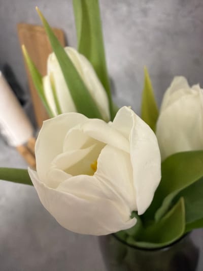 A close-up view taken indoors in the city of Ковалёво, Russia, presents a floral arrangement of three white tulips. The prominent tulip in the foreground is partially open, displaying soft white petals that transition to a creamy yellow center, where a yellow pistil is visible. Two additional white tulips, appearing more closed, are positioned in the background to the left and right. All tulips possess green stems and leaves. They are arranged in a dark, cylindrical vase, partially visible at the bottom of the frame. The background consists of a textured, light gray surface, likely a wall. Blurred elements are discernible in the upper left background, including a light brown rectangular object and a pale, elongated cylindrical item. No individuals are present.
