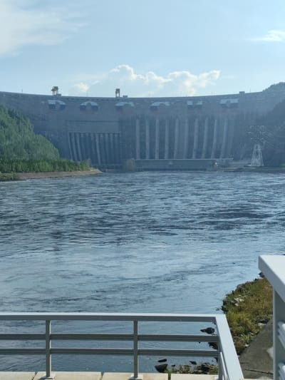 This image captures a wide view of the Sayano-Shushenskaya Hydroelectric Power Station, a colossal dam situated in Cheremushki, Russia. The imposing concrete structure dominates the background, stretching across the horizon with its massive spillways and crest, adorned with what appear to be stylized Russian flags. Tall towers and power lines are visible on the right side, suggesting the scale of electricity generation.
In the foreground, a calm, wide river flows, its surface rippled by gentle currents, reflecting the pale blue sky and scattered white clouds. The water's surface is a mix of dark blues and lighter streaks, hinting at the depth and movement within the river. The bank on the right is rocky and covered with dry grass. A white metal railing runs across the bottom of the frame, indicating a viewing platform or walkway, allowing visitors a vantage point to appreciate the grandeur of the dam and the surrounding landscape.
The scene is bathed in the soft light of what appears to be daytime, possibly late morning or early afternoon, given the bright sky. The weather seems fair with a few fluffy clouds dotting the sky. The lush green trees lining the left bank of the river contrast with the stern gray of the concrete dam. The overall impression is one of immense scale and human engineering, set against a natural, albeit controlled, environment. There is no visible text in the image.