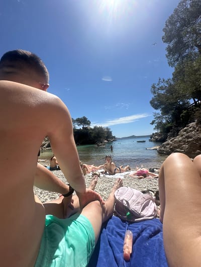The image shows a sunny beach scene in Peguera, Spain. The foreground features a person's shoulder, arm, and legs wearing green shorts, with another person's shoulder and arms. Behind them, several people are sunbathing on the pebble beach. There's a light pink backpack, a blue towel, and a bottle of lotion on the beach. In the background, there's the sea with a few boats, and rocky cliffs covered with trees. The sky is blue with a few clouds. A bird flies in the sky.