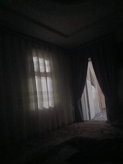 The image depicts a dimly lit room with heavy curtains drawn over a window. The curtains are a dark color and appear to be made of thick fabric, obscuring most of the light from outside. A sliver of light is visible through a gap in the curtains, illuminating a portion of a white, sheer curtain with a delicate leaf pattern. The room appears to be carpeted. To the right, a doorway is partially open, revealing another room or hallway beyond, also in shadow. The overall impression is one of privacy and subdued lighting, possibly indicating evening or a desire for darkness. The location is identified as Denov, Uzbekistan.