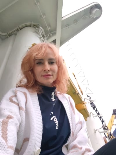 An outdoor, eye-level, medium shot shows a woman with peach-colored hair and makeup, wearing a white cardigan with peach-colored stripes over a navy blue shirt with white lettering. The woman is positioned in the lower-left of the frame. The background consists of white pipes, cables, and metallic structures, suggesting an industrial or maritime setting, possibly aboard a ship or in a port. The sky is overcast and white, with no visible details of Nikol'skaya Gora, Russia.