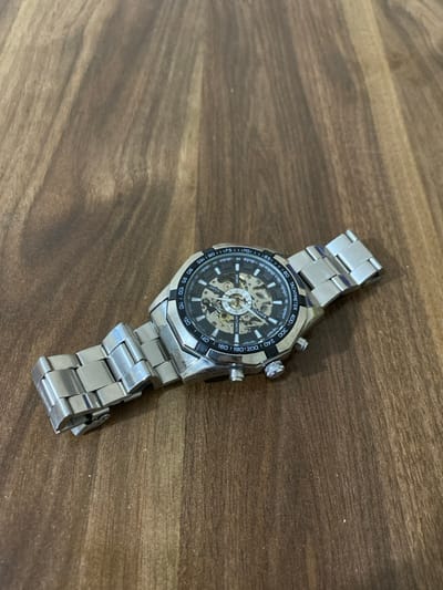 A silver-toned automatic wristwatch with a skeleton dial is depicted resting diagonally on a dark wooden surface. The watch features a prominent round face with visible internal gears and components, enclosed by a metallic bezel. A silver link bracelet is attached to the watch case, with the clasp end facing the bottom right.
The watch face is black with white hour markers and indices, along with a tachymeter scale around the outer edge. Visible text on the dial includes "TACHYMETER," numbers such as "400," "300," "240," "200," "180," "160," "140," "120," "110," "100," "95," and "90." The brand name "WANNEE" is visible on the upper part of the dial. Below the center, the words "SPORT" and "AUTOMATIC" are clearly legible, with "WATCH" also partially visible next to "SPORT."
The wooden surface, likely a table or floor, exhibits a distinct dark brown grain pattern. The lighting is diffused, suggesting an indoor setting without direct sunlight. The watch is the central and only subject, positioned from the upper left to the lower right of the frame. The scene is set in Abad Tukimachi, Uzbekistan.