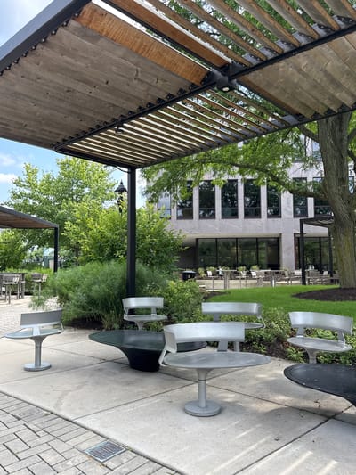 This is an outdoor seating area under a wooden pergola with metal supports. The area has a paved walkway and is furnished with several modern, minimalist silver tables and chairs, as well as a dark, conical table. In the background, there is a building with a light-colored granite facade and large, dark-framed windows. Green trees and lush bushes surround the seating area and the building. A bronze plaque is embedded in the paving stones in the lower right portion of the image. The plaque reads: "DOWNTOWN NAPERVILLE", "HISTORIC ILLINOIS", "PRESERVATION AWARD", "NAPERVILLE HERITAGE ASSOCIATION", "2019". This indicates the location is in downtown Naperville, Illinois, USA. No people are visible in the immediate seating area, though a person in a light-colored shirt can be seen walking in the distance on a path in front of the building. The sky is partly cloudy.