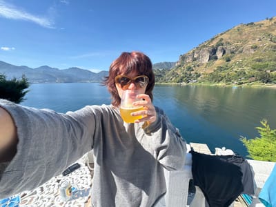 The image captures a selfie taken from a balcony or terrace overlooking Lake Atitlán in San Pablo La Laguna, Guatemala, under a clear blue sky. A female subject with reddish-brown hair and brown sunglasses, wearing a grey long-sleeved top, is centrally positioned, bringing a clear, stemless glass containing an amber liquid to her mouth with her right hand. A silver ring is visible on her right ring finger, and her fingernails are painted yellow. Her left arm is extended forward, holding the camera. In the immediate foreground, to the left of the subject, a white, ornate patterned surface or table holds two aluminum cans. To the right, a black swimsuit is draped over a white railing. The background features a vast expanse of dark blue lake water, with ripples on its surface, flanked by steep, verdant mountains. The mountains to the right exhibit rocky outcrops and scattered structures along their slopes. The sunlight is bright, casting distinct shadows, indicating midday or early afternoon.