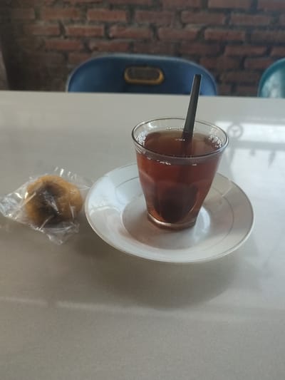 A glass of iced tea with a spoon is placed on a saucer on a light-colored table. To the left of the glass, a small, round pastry wrapped in clear plastic is visible. In the background, a brick wall and two chairs can be seen. The scene appears to be set in Pantepisang, Indonesia, possibly in a cafe or restaurant.
