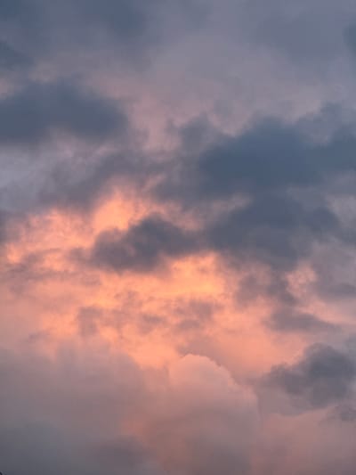 The image captures a sky filled with dramatic cloud formations, bathed in the soft, warm light of either sunrise or sunset. Dominant colors include various shades of greyish-blue and purple in the shadowed areas, contrasting sharply with luminous patches of peach-orange and pink. These warm hues are most prominent on the right side and center of the frame, where the sunlight appears to be directly hitting the clouds, creating a vibrant glow.
The clouds display a varied texture, ranging from thick, dense masses that block the light to more ethereal, wispy sections. Some formations show clear definition and layering, while others appear more diffused and blended. The overall scene suggests a partly cloudy sky, with the sun low on the horizon, illuminating the atmospheric conditions from below.
There are no discernible people, objects, or specific events taking place, focusing entirely on the natural beauty of the sky. The image conveys a serene yet powerful natural moment. The location is Fécamp, France, although no specific landmarks or geographical features are visible within the frame. There is no visible text in the image.
