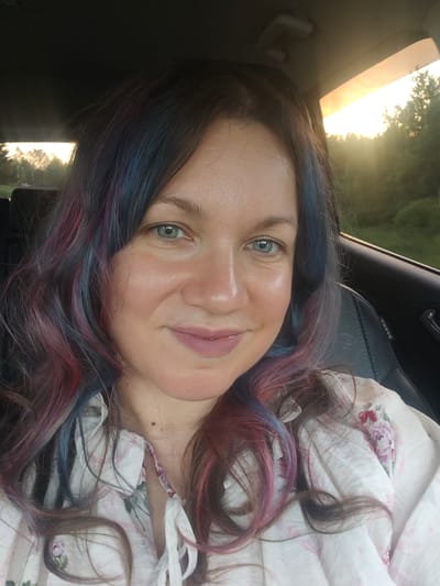 A selfie of a woman with fair skin, blue eyes, and multicolored hair (dark brown with streaks of blue, red, and pink). She is wearing a light-colored floral print blouse. The background appears to be the interior of a car, with a glimpse of a wooded area and a sunset through the window. It is not possible to confirm if it is in the city of Ильинское, Russia.