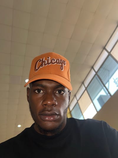 A close-up, low-angle shot depicts a dark-skinned man wearing an orange baseball cap with "Chicago" embroidered on the front. He is also wearing a black shirt. The background consists of a ceiling with a grid of rectangular light panels. The location is not in the city of Kwi, Nigeria.