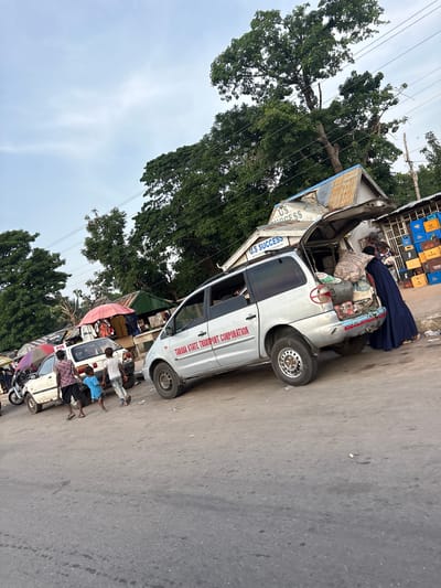 The image depicts a busy street scene in Gulum, Nigeria, with a focus on a silver minivan packed with goods. The minivan's rear hatch is open, revealing several large sacks and other items being loaded or unloaded. A woman in a long navy blue dress stands at the back of the van, interacting with the contents. In front of the van, a white car is parked, and several people, including two young boys and an adult male, are walking nearby. A red umbrella is visible behind the white car, along with some market stalls. The environment suggests a marketplace with various vendors and customers. The sky is overcast, and large trees line the background. Signs on the building behind the van read "U.S SUCCESS" and "U.S SUCCESS SHOP". The road surface is asphalt with some debris.