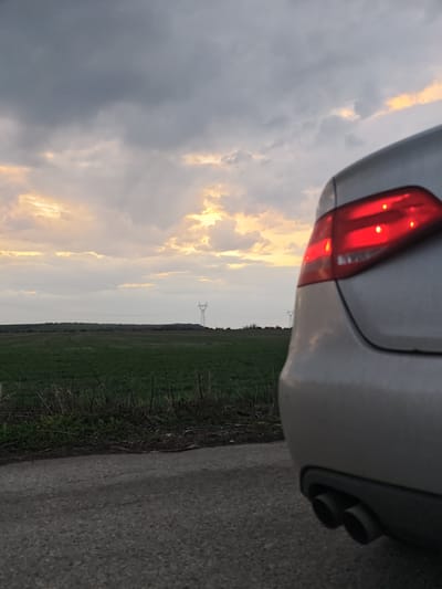 The scene depicts the rear quarter of a light-colored sedan, possibly silver, parked on an asphalt road in a rural setting within Gorni Tsibar, Bulgaria. The vehicle's red taillights are illuminated, and a dual exhaust system is visible on its lower right side. To the left of the road, a strip of dry, dark vegetation borders an expansive green agricultural field. In the mid-ground, at least two metal lattice power pylons are visible against the horizon, with power lines extending between them. The sky is largely overcast with dark grey clouds, punctuated by patches of orange and yellow light, suggesting either sunrise or sunset. No individuals are present in the image.