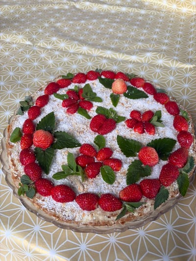 A round, multi-layered cake is decorated with strawberries and mint leaves, and dusted with powdered sugar and shredded coconut. The cake sits on a clear glass cake stand, which is placed on a yellow tablecloth with a geometric pattern of white stars. The scene appears to be indoors, possibly in a home setting, with natural light illuminating the cake. There is no indication of the city of Субханкулово, Russia in the image.