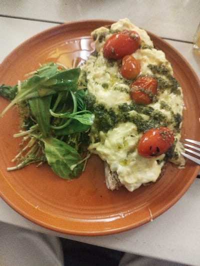 An orange plate presents a meal of eggs, tomatoes, pesto, and greens. The creamy eggs, dotted with green pesto, sit atop bread and are garnished with roasted red tomatoes. A cluster of fresh, green leaves rests to the left of the eggs, adding a vibrant touch to the dish. A fork is visible beside the eggs.
