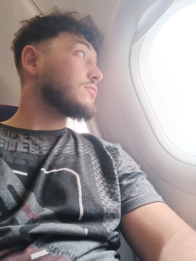 The image depicts the interior of an aircraft. One male individual is present, positioned in profile facing the right side of the frame. He has dark, curly hair with a faded haircut on the sides, and a dark beard and mustache. He is wearing a short-sleeved, dark gray t-shirt with a patterned design on the upper half, featuring horizontal lines and a grid-like texture, along with white and reddish graphic elements on the front. The individual is looking upward and to the right, out of an oval-shaped aircraft window that shows an intensely bright, featureless white exterior, indicative of daylight or clouds. Part of a dark red or purple seatback is visible behind his left shoulder. The aircraft cabin ceiling is visible at the top. The specific location within the city of Saint-Louis, France, cannot be determined from this image, as it shows an airplane interior, which could be in flight or at any airport.