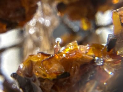 The image is a close-up, out-of-focus photograph of what appears to be amber resinous material. The material is a translucent, deep orange-brown color with lighter, almost white, irregular inclusions. There are also areas of lighter yellow and brown. The surface of the material is textured, with some parts appearing smooth and glossy, while others are rough and crystalline. Small, bright highlights suggest a light source reflecting off the surface. The overall impression is of a natural, perhaps fossilized, substance. The location within the city of Biržai, Lithuania is not discernible from the image itself; it is a close-up macro shot of an object.