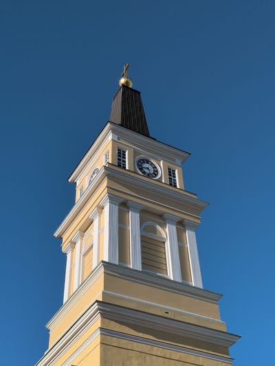The image shows a close-up, low-angle view of the yellow and white clock tower of Oulu Cathedral, located in Oulu, Finland. The sky is a clear, deep blue. The tower has a dark, pointed spire topped with a golden orb and a cross. The clock face is visible on one side of the tower, with Roman numerals and ornate hands. The architectural details of the tower, including columns and arched windows, are clearly depicted. The lighting suggests it is daytime, with the sun illuminating the building. There are no people visible in the image.