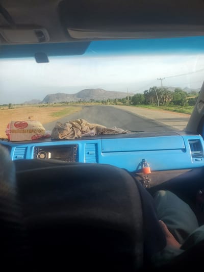 A first-person perspective shot from inside a vehicle reveals a dashboard, a windshield, and a view of the road ahead. The dashboard is a bright blue color and features a radio and vents. A red padlock is attached to the dashboard. On the dashboard, in front of the radio, sit two items: a white package wrapped in plastic with red and black lettering that reads "NEW NATIONAL," and a pile of beige fabric.

Outside the vehicle, a paved road stretches into the distance, flanked by a dry, dusty landscape. In the background, rolling hills and mountains are visible under a hazy sky. Trees and vegetation dot the sides of the road, and power lines with poles run parallel to the path. The scene appears to be set in a rural or semi-arid environment. The exact location is specified as Karihe, Nigeria.