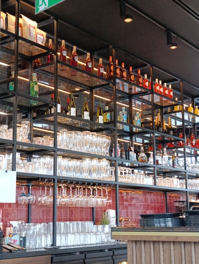 The image depicts a well-stocked bar shelf in a commercial establishment, likely a restaurant or pub, located in Mysiadło, Poland. The shelves are made of black metal and are filled with various bottles of liquor, wine, and other beverages. Several rows of glassware, including wine glasses and tumblers, are neatly stacked on lower shelves. The background features a red tiled wall. The lighting in the establishment appears to be ambient, with some spotlights casting a warm glow. The overall impression is one of a clean and organized bar area, ready for service. No people are visible in the image.