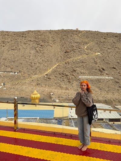 A young woman with curly brown hair and an orange headscarf stands on a raised platform in Chumkar, India. She is wearing a tan sweatshirt and light blue jeans, and is barefoot. She holds her hands clasped in front of her chest and carries a black tote bag with white lettering. The platform has a red and yellow striped pattern. Behind her, a steep, rocky hillside is visible, with a winding yellow path leading towards the top. Large white letters spelling "DHAN GURU NANAK DEV JI" are visible on the hillside. A golden dome structure is also present on the hillside, and a metal railing runs along the edge of the platform. The sky is overcast.