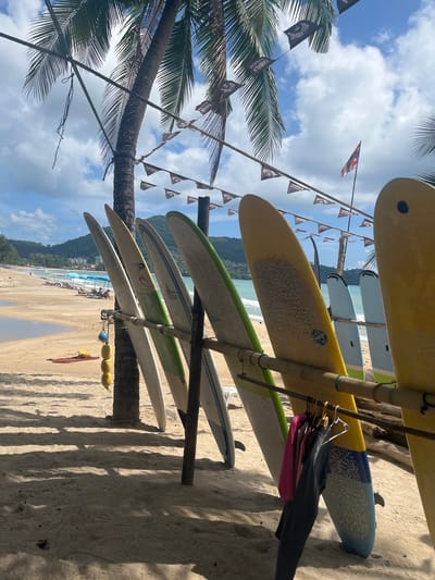 In Ban Bang Thao, Thailand, a row of surfboards stands upright on a sandy beach, leaning against a bamboo rack. The surfboards vary in color, with some displaying yellow, green, and white hues. Clothing items, including a pink shirt and dark pants, hang on hangers attached to the rack below the surfboards. In the background, a tropical beach scene unfolds with palm trees, beach umbrellas, lounge chairs, and the ocean under a partly cloudy sky. A small flag is visible on a pole in the distance. The ground is covered in sand, with shadows cast by palm fronds.