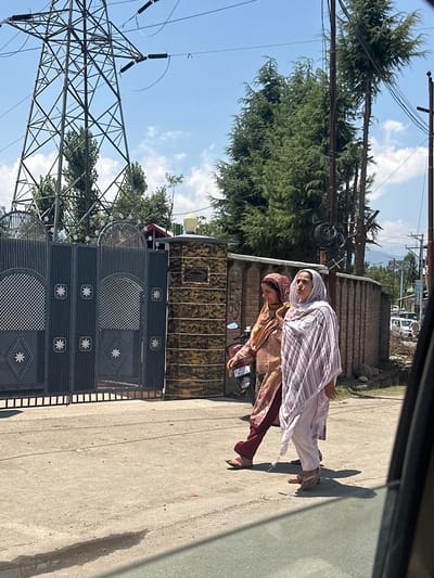 Two women, one wearing a burgundy and gold patterned salwar kameez and a maroon headscarf, the other wearing a pale pink and white striped shalwar kameez and a pale pink headscarf, walk on a paved road in Vessu, India. The woman on the left is partially obscured by the woman on the right. A motorcycle with license plate JK03 M9056 is parked near them. In the background, a tall, latticed metal electricity pylon stands against a blue sky with scattered clouds. Behind the women, a brick wall encloses a property with a large, decorative metal gate. Trees with dense green foliage are visible behind the pylon and the wall. Further in the background, more trees and buildings are present. The ground is a light gray asphalt with some debris and a wet patch. A portion of a dark-colored vehicle is visible in the foreground, obscuring the lower right of the frame.