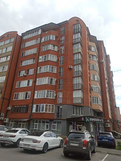 In Zavodskoy, Russia, a modern, multi-story brick apartment building dominates the scene under a cloudy sky. The building features numerous balconies with white-framed windows and dark metal railings, along with external air conditioning units attached to the brickwork. The ground level of the building houses commercial spaces, including a store labeled "Kinder" with Cyrillic text, and an adjacent business with large glass windows. In front of the building, a row of parked cars, including a white Genesis sedan, a white Lada sedan, a dark gray SUV, and a blue Hyundai SUV, are visible on a paved surface. Power lines are strung across the upper right portion of the image.