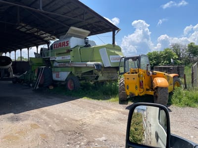An outdoor agricultural scene in Gorni Bogrov, Bulgaria, is depicted on a bright, sunny day with a clear blue sky adorned with large, fluffy white cumulus clouds.
In the foreground, a large green and white Claas combine harvester is partially parked under an open-sided shed with a corrugated metal roof, casting a shadow over the foreground ground. Visible text on the combine includes "CLAAS," "MAXI," and "DOMINATOR 108 SL." A red ladder leans against the rear wheel of the combine. To the right of the combine, a yellow JCB telehandler is parked on the unpaved ground, which is a mix of dirt and gravel. Several muddy puddles are present on the ground, indicating recent wet conditions.
Beyond the vehicles, green grass and weeds grow around the machinery and at the edges of the shed. Further in the background, a line of lush green trees and shrubs is visible under the bright sky. The shed itself has a visible metal framework supporting the roof, with glimpses of the sky through gaps in the structure.
The image is taken from the perspective of someone inside a vehicle, with a side-view mirror occupying the bottom right corner of the frame. The mirror reflects a portion of a white truck or van and some green foliage. No people are visible in the scene, and the heavy machinery appears to be stationary, possibly stored or awaiting maintenance.