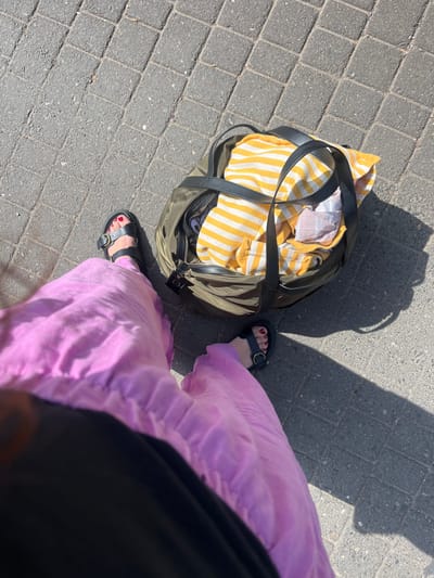 The image depicts a high-angle view of a person standing outdoors on a sunny day in Bad Sobernheim, Germany. The subject's lower torso and legs are visible, wearing loose, light purple trousers or a skirt and black sandals with a gold-colored buckle on each foot. The person's toenails are painted red. To the right of the feet, an olive green or dark khaki duffel bag with black handles rests on the paved ground. The bag is partially open, revealing a yellow and white striped fabric and other lighter-colored materials inside. The ground is a paved surface composed of rectangular grey blocks laid in a herringbone pattern. Strong sunlight casts distinct shadows of the person and the bag onto the pavement, indicating the scene is brightly lit.