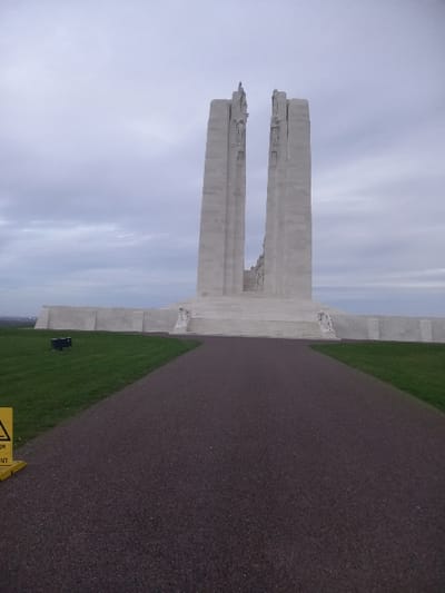 A tall, weathered white monument with two vertical columns stands against a cloudy sky. A pathway leads directly towards the structure, flanked by green grass. The monument shows signs of age and weathering. The path and grass are neatly maintained, creating a solemn and respectful atmosphere. A small yellow sign is visible in the foreground. The scene conveys a sense of history and remembrance.