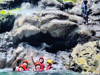 This image depicts a water-based recreational scene within a natural rock formation in Desa Pagandaran, Indonesia. Five individuals are partially submerged in clear, light blue-green water in the lower frame. All five wear orange life vests and orange or yellow helmets. Two individuals on the left are smiling; one extends two fingers in a 'peace' gesture, while the other raises a hand in a wave. In the upper right quadrant, one individual stands barefoot on a rock ledge, clad in a dark full-body wetsuit and a light green/yellow life vest. The background is dominated by a rugged, multi-layered rock face exhibiting grey, beige, and brown tones. Portions of the rock are covered with green moss, and visible water cascades down sections, forming stalactite-like structures. The environment is naturally lit, highlighting the textured rock surfaces and the water below.