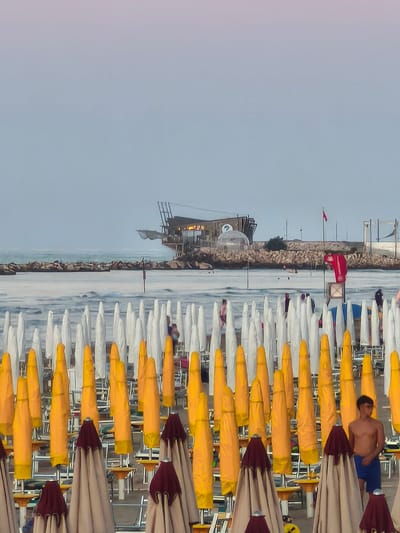 In San Salvo, Italy, a beach scene is depicted under a pale, hazy sky with hints of pink and purple at the horizon. Rows of closed beach umbrellas are lined up on the sand, predominantly yellow and white, with some beige and dark red umbrellas in the foreground. The umbrellas are organized in neat rows, suggesting a resort or public beach setting. In the middle ground, a man is visible standing between two umbrellas, wearing blue shorts. Further back, in the sea, a structure resembling a fishing platform or restaurant is built on stilts above the water, with additional structures and a dome-like enclosure visible on a rocky outcrop. Several people are seen on the beach and near the water, though their details are indistinct. The sea appears calm, with a rocky breakwater extending into the water. The overall atmosphere is that of a quiet, late afternoon or early evening on the coast.
