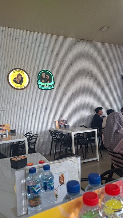 This is a photograph of the interior of a restaurant in Sigli, Indonesia. The restaurant features a wall with vertical corrugated panels, creating a textured background. Two illuminated signs are visible on the wall. The sign on the left is circular and yellow, with an image of a drink and text in Indonesian. The sign on the right is green and depicts two figures, with text in Indonesian.
Several tables with white tops and black chairs are arranged in the dining area. Some tables are set with bottles of water and napkins. In the background, several patrons are seated at tables, engaged in conversation or dining. One person is wearing a black shirt, and another is wearing a patterned scarf.
In the foreground, the bottom portion of the image shows several bottles of water with blue and red caps on a table, suggesting a recently served meal or beverages. The overall ambiance appears casual and social, typical of a local eatery. The lighting is bright, suggesting it is daytime.