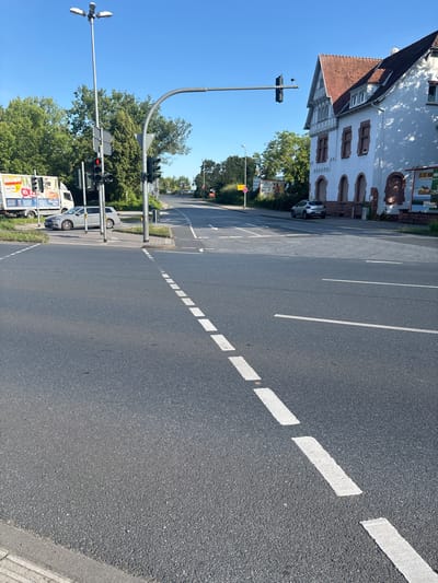 The image depicts a street scene in Groß-Gerau, Germany, on a clear, sunny day. The foreground is dominated by an asphalt road with white dashed lines indicating a lane or pedestrian crossing. To the right, a light-colored car is parked at an angle to the road. In the middle ground, a modern traffic light with red and yellow signals is visible, mounted on a tall metal pole. Adjacent to it is a building with a red tiled roof and white walls, featuring arched windows and decorative architectural elements. A delivery truck is parked to the left, with advertisements on its side. Trees line the background, and more traffic lights and street signs are present further down the road. The environment appears to be an intersection or a busy street in a town or city.