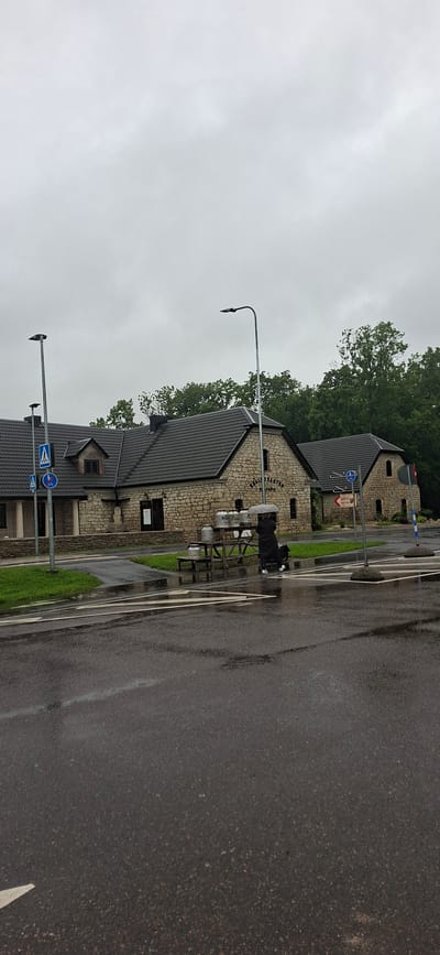 The image depicts an outdoor scene on a cloudy day, likely in Kaali, Estonia. In the foreground, a wet asphalt road with white lane markings is visible. A pedestrian crossing is marked on the road. To the left of the road, there is a grassy area with a stone wall. Beyond the wall, a stone building with a dark, pitched roof is situated. The building has several windows and at least two doorways.
To the left of the building, a lamppost stands near a pedestrian crossing sign. In front of the building, there is a wooden picnic table laden with several large, metallic milk churns. A person wearing a dark coat and holding a clear umbrella stands near the table, seemingly interacting with the churns. Another lamppost is visible to the right of the building. In the background, lush green trees form a dense line. Further to the right, another similar stone building is partially visible, and a red and white sign on a post is present. The overall impression is of a rural or semi-rural setting, possibly a farm or a visitor center, under overcast conditions.