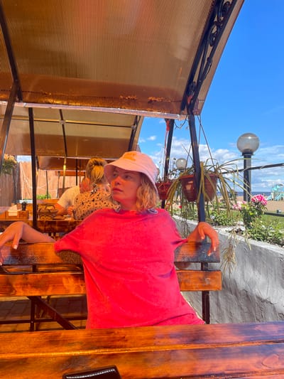 The image shows a young woman with blonde hair wearing a pink bucket hat and a bright pink t-shirt, sitting on a wooden bench under an orange translucent awning. She is looking to her right with a contemplative expression. Behind her, other people are visible at tables, and plants are hanging in pots. In the background, there is a gray concrete wall, a street lamp, and a view of the water and a playground. The scene appears to be an outdoor seating area of a cafe or restaurant. The image is likely taken in the city of Belozersk, Russia, given the context.