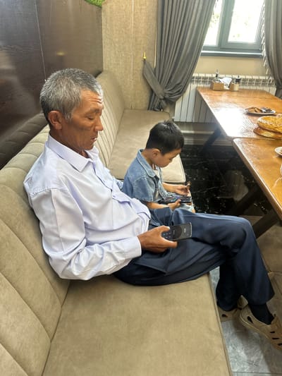 In Ынтымак, Kyrgyzstan, an elderly man with graying hair and a light blue button-down shirt sits on a beige couch, holding a smartphone. Beside him, a young boy in a denim shirt looks at his own phone. The man's right hand rests on the phone, with his thumb on the screen. The couch is upholstered in a textured beige fabric with vertical stitching. In the background, a wooden table is visible, set with plates of food and a salt and pepper shaker. A window with grey curtains is also present. The flooring appears to be black and white marble.