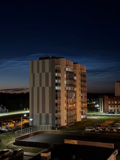 The image is a nighttime, slightly low-angle, wide shot of a modern apartment building in Tsiolkovsky, Russia. The building has a gray and white checkered facade with numerous windows, many of which are lit from within, suggesting occupancy. To the right of the apartment building, a curving, elevated walkway with white railings and stairs is visible, leading to a dark, paved area that appears to be a basketball court with a red hoop. Beyond the apartment building, a parking lot filled with cars is illuminated by streetlights. The sky is a deep blue, transitioning to a lighter hue near the horizon, with wispy clouds faintly visible. The overall setting is an urban residential area at dusk or night. There is no visible text in the image.