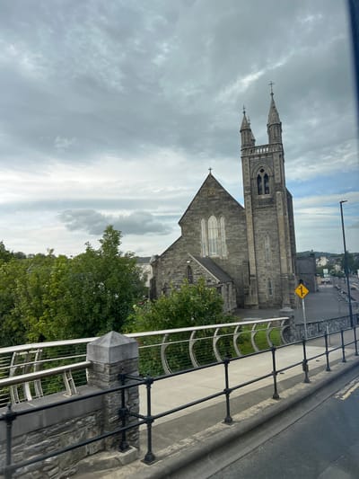 The image captures a view of St. Mary's Church in Ballybofey, County Donegal, Ireland, as seen from a bridge. The church is a prominent stone structure with a tall tower featuring spires and crosses, a pitched roof, and arched windows. Adjacent to the church are lush green trees. In the foreground, a pedestrian walkway is visible, bordered by a modern railing system made of stone and metal. The walkway leads to a street, with a yellow diamond-shaped traffic sign for a right turn visible. The sky is overcast with a mix of clouds. The scene suggests a partly cloudy day.