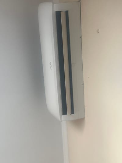 The image shows a white split-system air conditioner mounted on a light-colored wall. The air conditioner has a prominent vent with dark slats. A subtle, silver logo, appearing as two interconnected 'W' shapes, is visible on the front panel of the unit. The wall to the right of the air conditioner has a small, circular indentation and a faint dark mark. The lighting suggests an indoor environment, possibly a room in a building located in Lebedinovka, Kyrgyzstan, though this cannot be definitively confirmed from the image alone. The image is oriented vertically, with the air conditioner running from bottom to top.