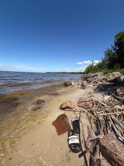 A pair of black Givenchy Paris slides rests on a sandy beach in Lebedivka, Ukraine. The slides are positioned on the sand near a collection of driftwood and large rocks. The scene depicts a sunny day with a clear blue sky and calm water lapping at the shore. The water is a murky brown color, and the beach is composed of fine sand and scattered stones. The background shows a line of trees and vegetation along the horizon.