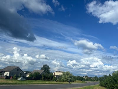 This is a wide-angle, eye-level shot of a semi-rural landscape in Грозаўскі сельскі Савет, Belarus, dominated by a dynamic sky. A clear blue sky occupies the upper two-thirds of the frame, streaked with wispy cirrus clouds and punctuated by larger, puffy cumulus clouds, some of which are tinged with grey, suggesting recent or impending precipitation. The lower third of the image depicts a road running diagonally from the foreground towards the middle ground, bordered by a grassy verge.
On the left side of the road, a row of houses with grey, pitched roofs are visible, interspersed with green trees and fences. One house is lighter in color with a dark-sloped roof and a small white extension on the left. A white sedan is parked in front of this house on the grass. To the right of these houses, the landscape continues with more trees and buildings, including a lighter-colored structure with a darker pitched roof further back. A single person, wearing dark clothing, is walking on the right side of the road in the middle distance. Dense green foliage lines the right side of the road, extending into the background where more distant trees and buildings can be discerned against the horizon. The overall impression is one of a bright, overcast day in a residential area outside a major city.