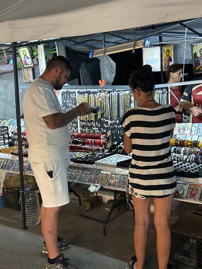 The image depicts an outdoor market stall, likely in the city of Stob, Bulgaria, given the religious iconography visible. In the foreground, a man with dark hair and a beard, wearing a white t-shirt and white shorts, is looking at a mobile phone in his hands. To his right, a woman with dark hair tied up in a bun, wearing a black and white striped t-shirt dress, is also looking at the merchandise displayed on the stall. The stall is laden with various items, including what appear to be religious icons or prints on the lower shelves, and numerous bracelets and necklaces on display behind them. A large plastic bottle of water hangs from a support structure to the left of the man. The background is dark, suggesting it is evening or nighttime, with the stall illuminated by artificial light. Another woman, with brown hair, is partially visible behind the main subjects, looking towards the right of the frame. The ground appears to be asphalt or a similar surface.
